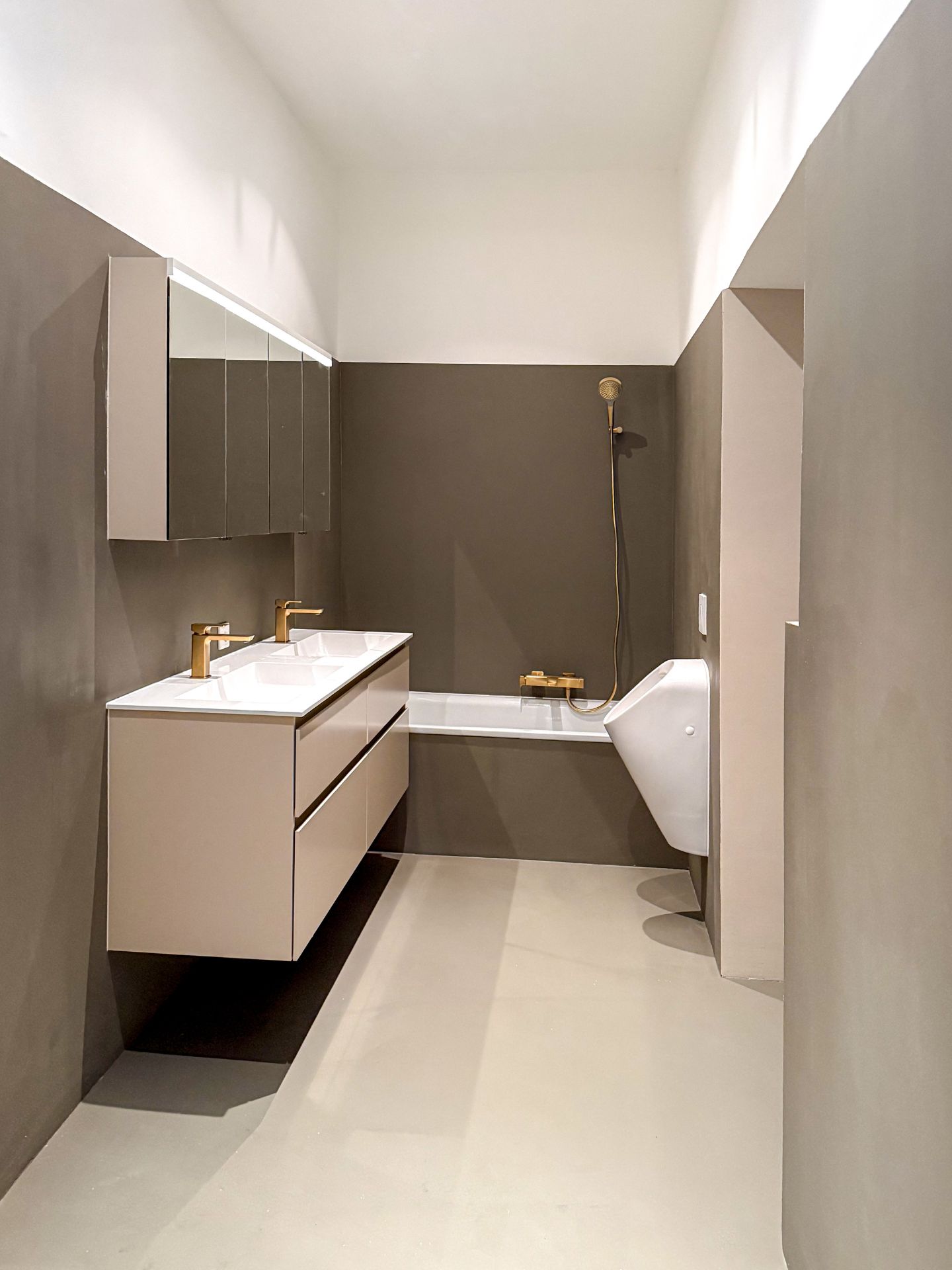 Modernes Badezimmer nach Komplettsanierung mit fugenlosem Mikrozement auf Boden und Wänden, eleganten goldenen Armaturen, Doppelwaschtisch mit Spiegelverbau sowie Badewanne und Urinal im minimalistischen Design.